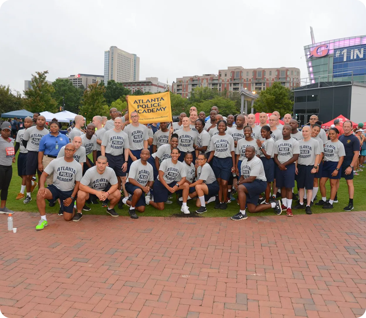 Group of Atlanta Police Academy trainees in gray t-shirts and navy shorts posing together outdoors, with one holding a yellow Atlanta Police Academy banner.