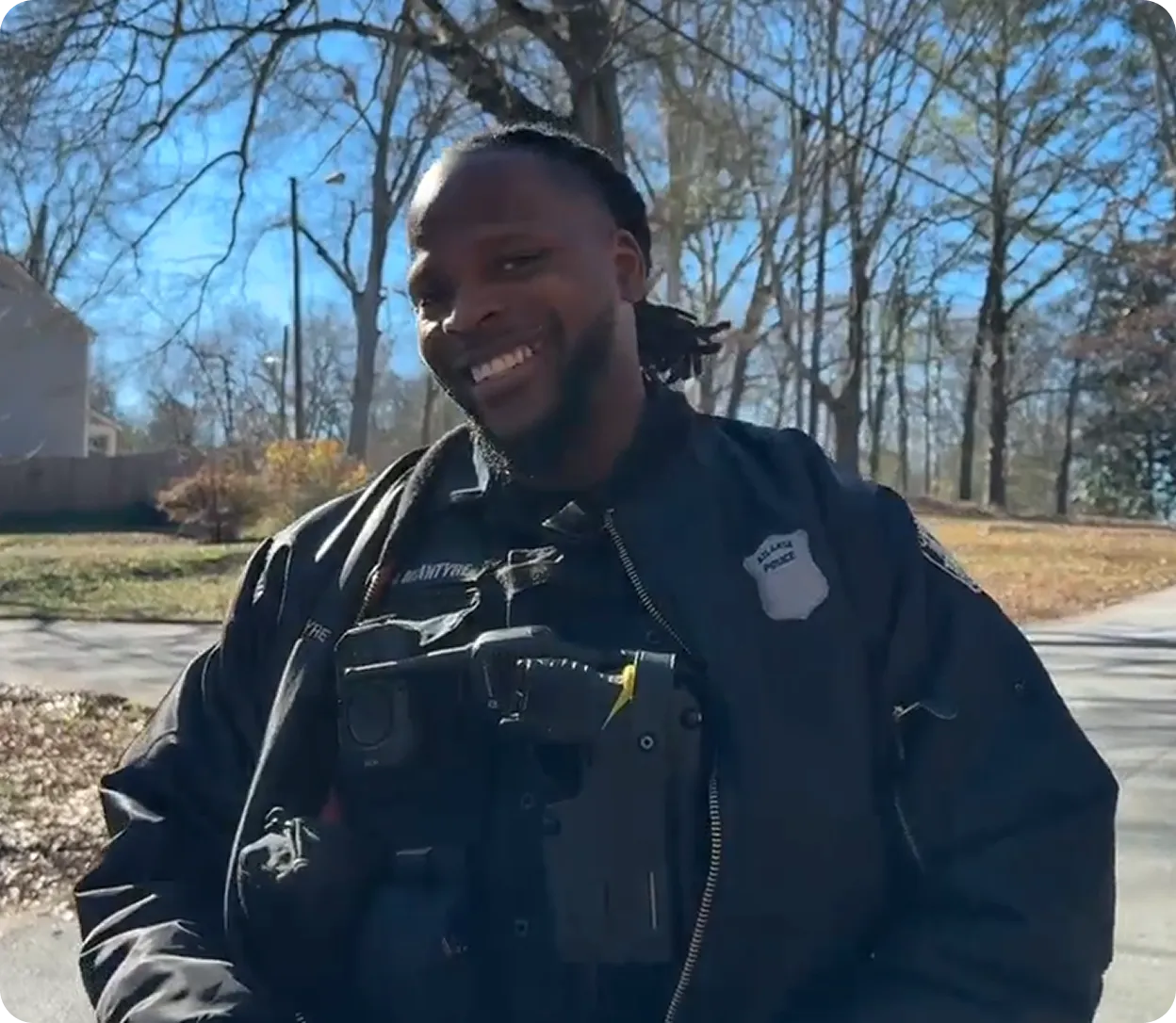 Smiling Black police officer with dreadlocks outdoors in a neighborhood with trees and houses in the background.