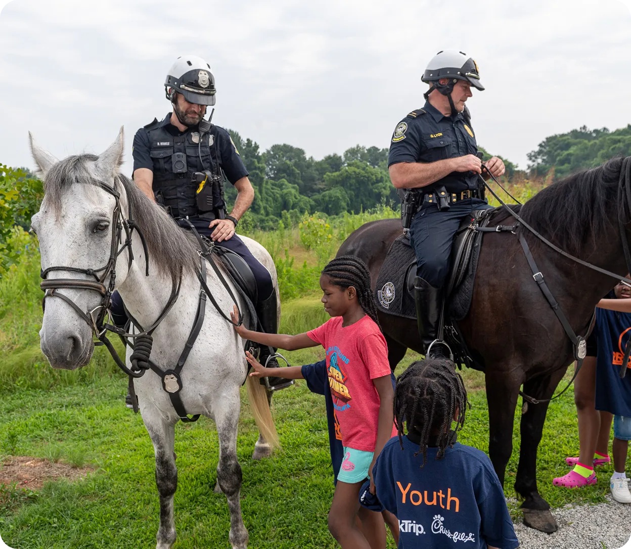 Two police officers on horseback interacting with children petting the horses outdoors on grass.