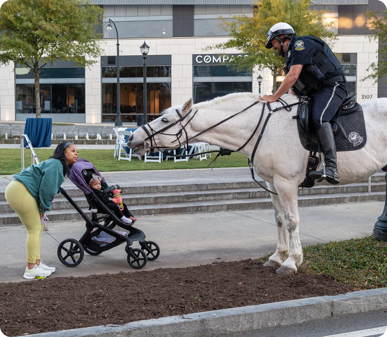 Woman leaning down to interact with a baby in a stroller as a police officer on a white horse reaches towards them.