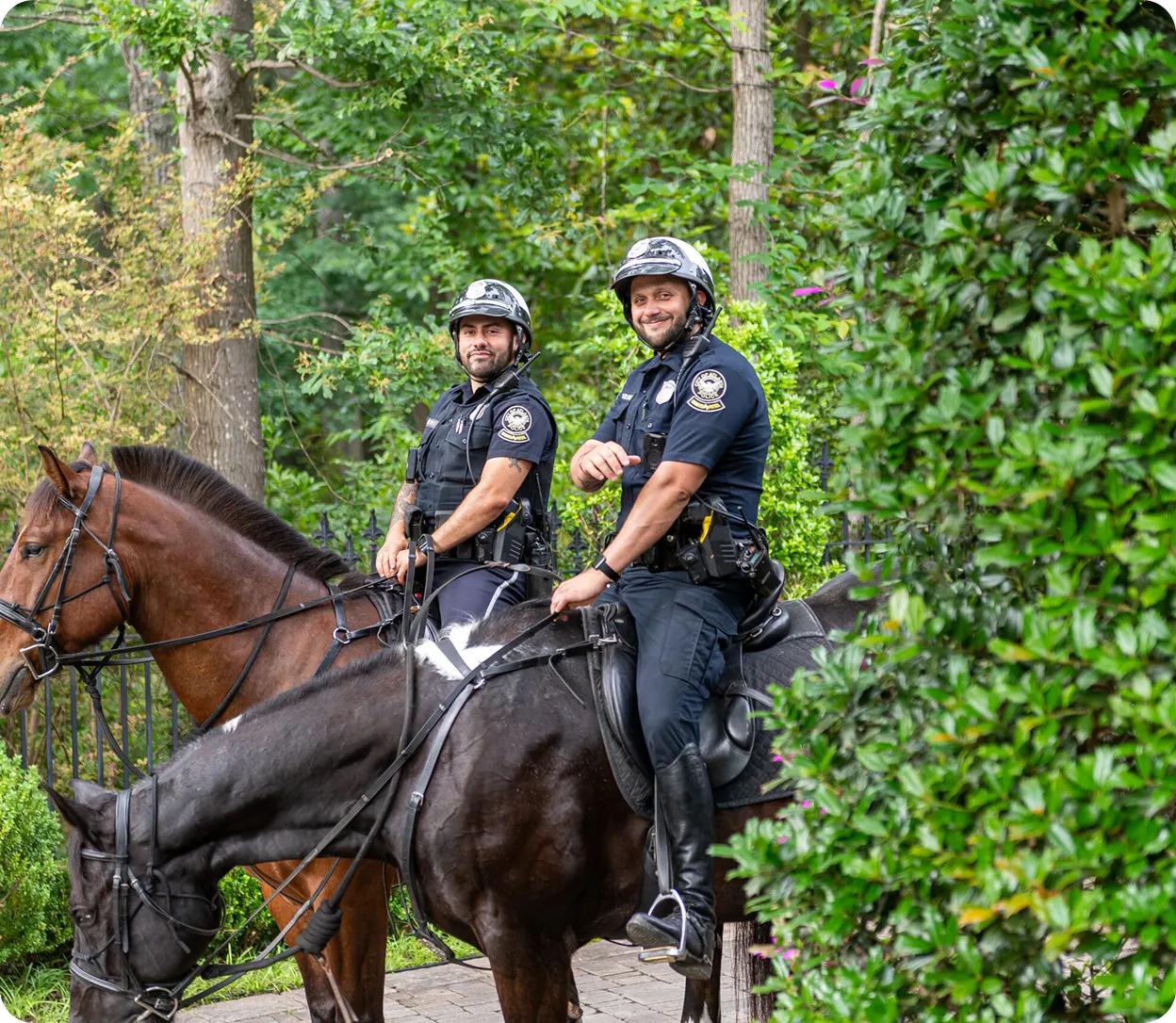 Two police officers in uniform and helmets sitting on brown and black horses in a leafy outdoor setting.