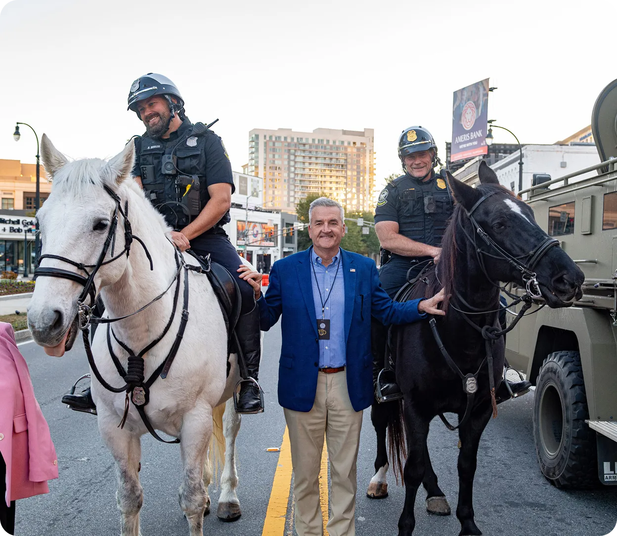 Two police officers in uniform on horseback smiling with a man in a blue blazer standing between the horses on a city street.