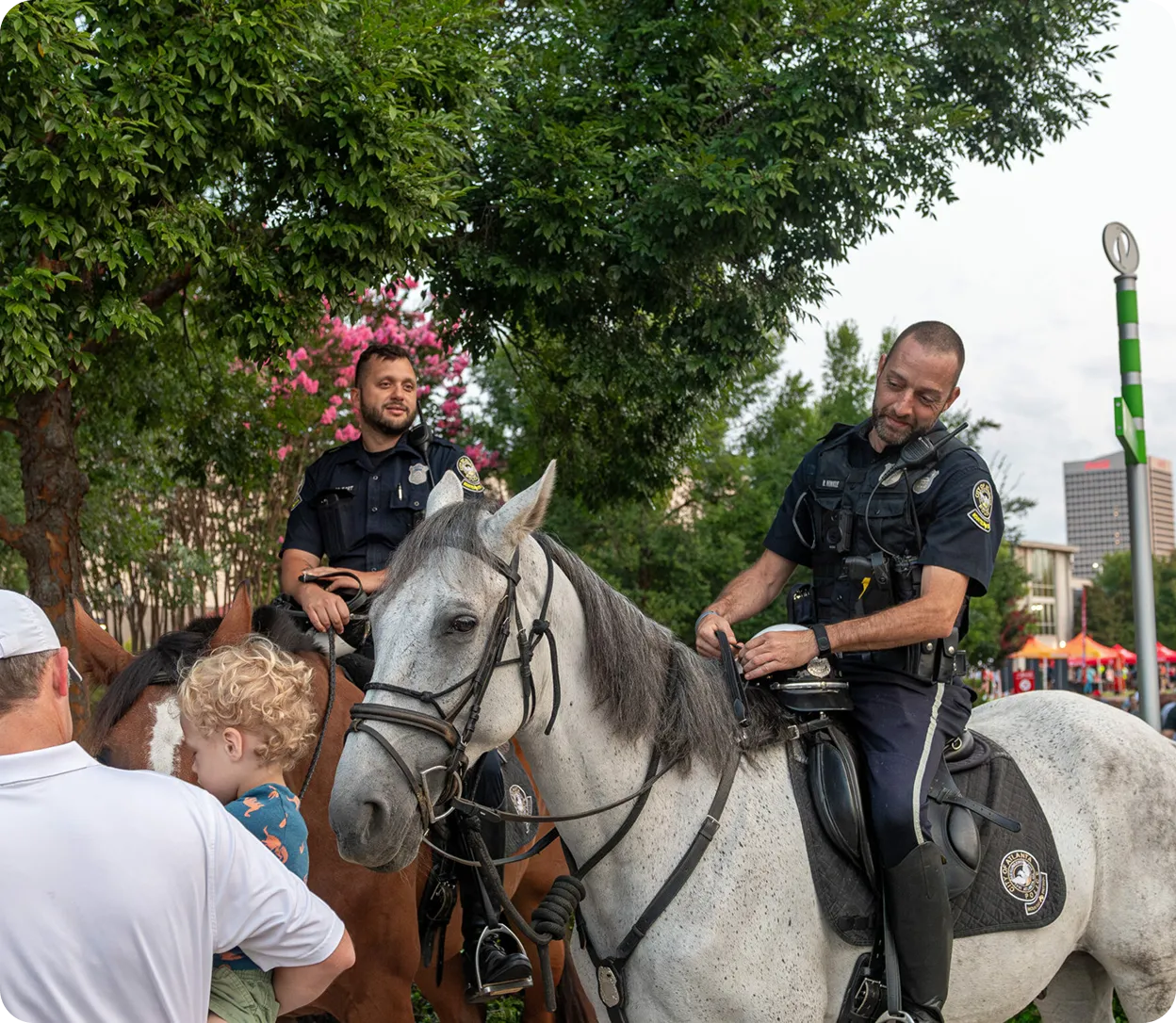 Two mounted police officers on horses interacting with a man holding a child in an outdoor urban setting with trees and buildings in the background.
