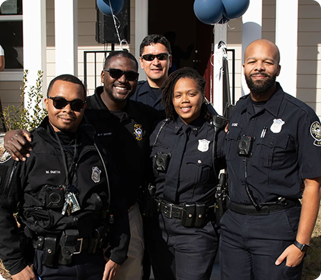 Five smiling police officers standing close together outdoors, three in uniform and two in black jackets and sunglasses.