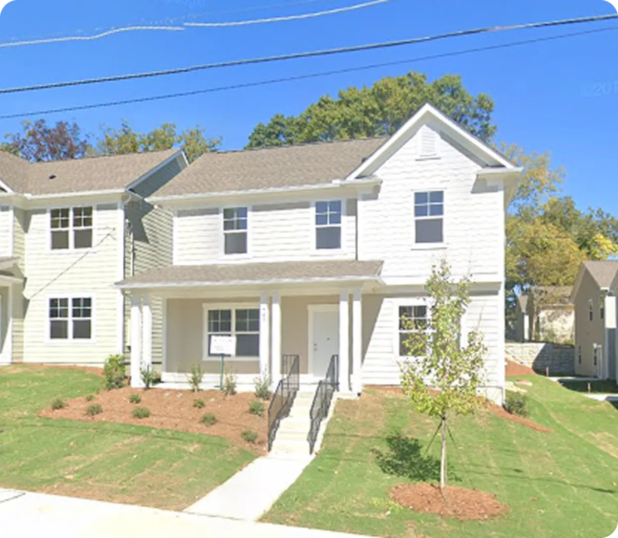 Two-story white house with a front porch, stairs, and newly planted landscaping on a sunny day.