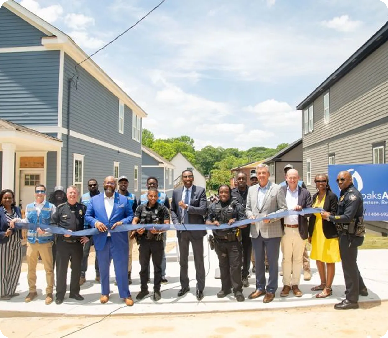 Group of people including police officers and city officials holding a blue ribbon at a ribbon-cutting event in a residential neighborhood.