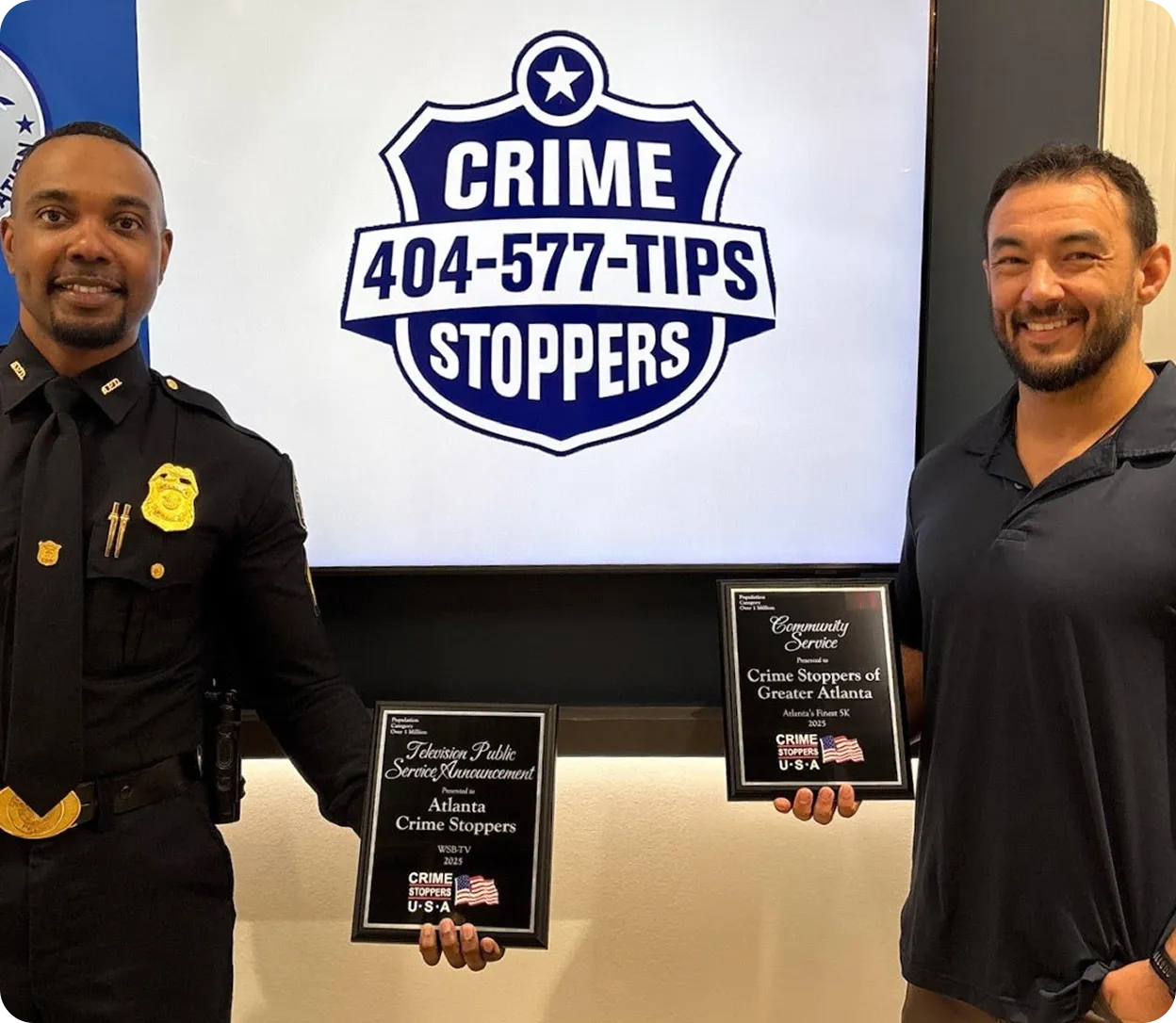 Two men holding plaques in front of a screen displaying the Crime Stoppers logo and tip line number 404-577-TIPS.