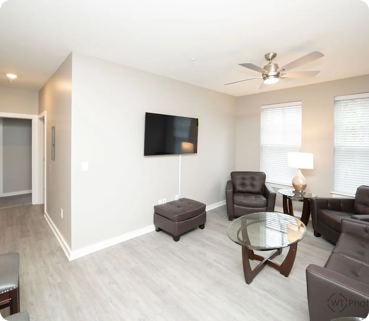 Modern living room with gray leather chairs and ottoman, glass coffee table, mounted flat-screen TV, ceiling fan, and a lamp on a side table near two windows with blinds.