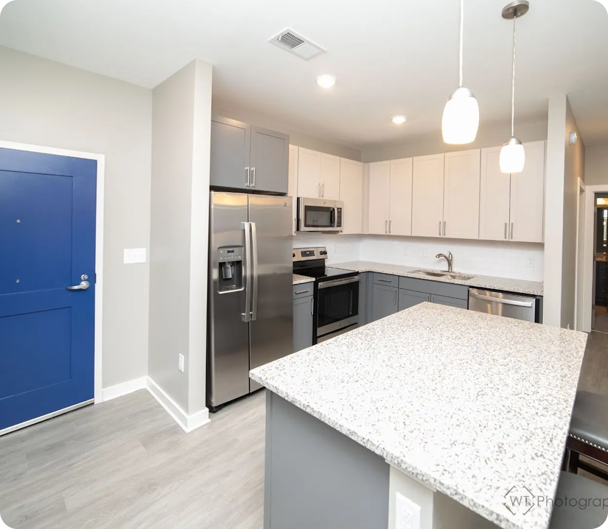 Modern kitchen with gray and white cabinets, stainless steel appliances, granite countertop island, and blue entry door.