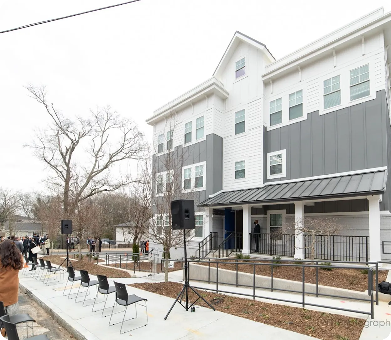 Outdoor event setup with black chairs arranged in rows and speakers in front of a modern three-story building painted white and gray.