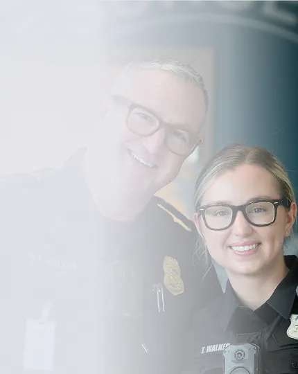 Two smiling police officers wearing glasses and uniforms, one male and one female, standing close together indoors.