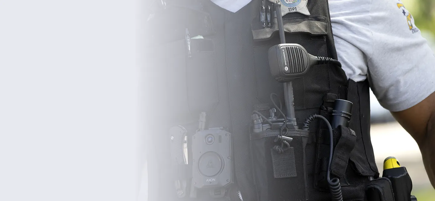 Close-up of a police officer's tactical vest showing a Motorola radio microphone, Axon body camera, flashlight, and other equipment attached to the vest.