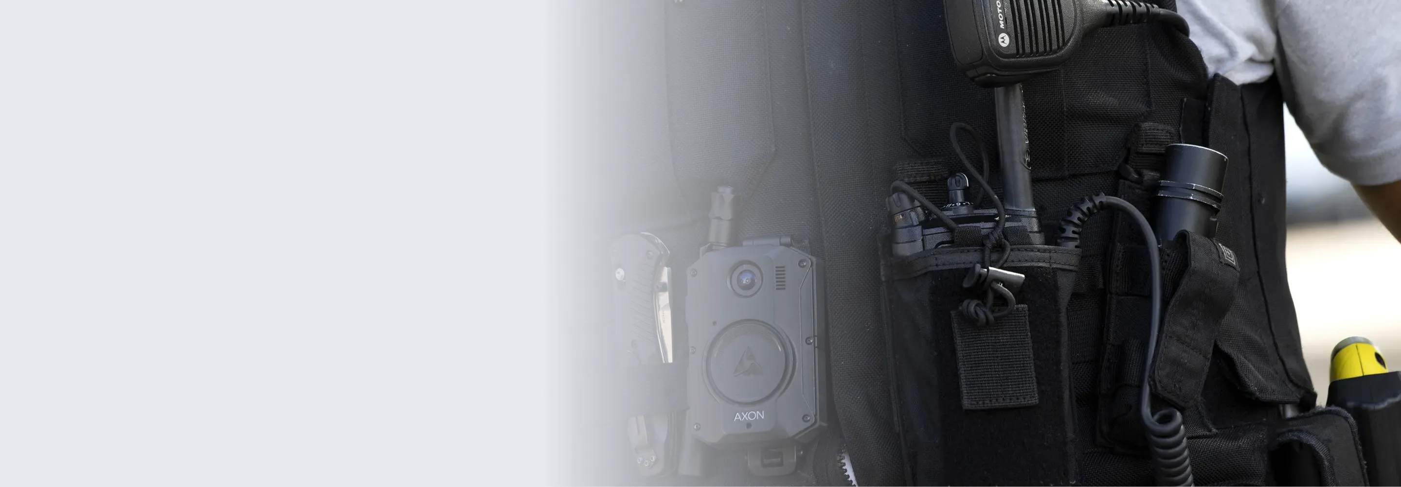 Close-up of a police officer's black tactical vest equipped with a body camera labeled AXON, a radio microphone, and other gear including a flashlight and a baton.