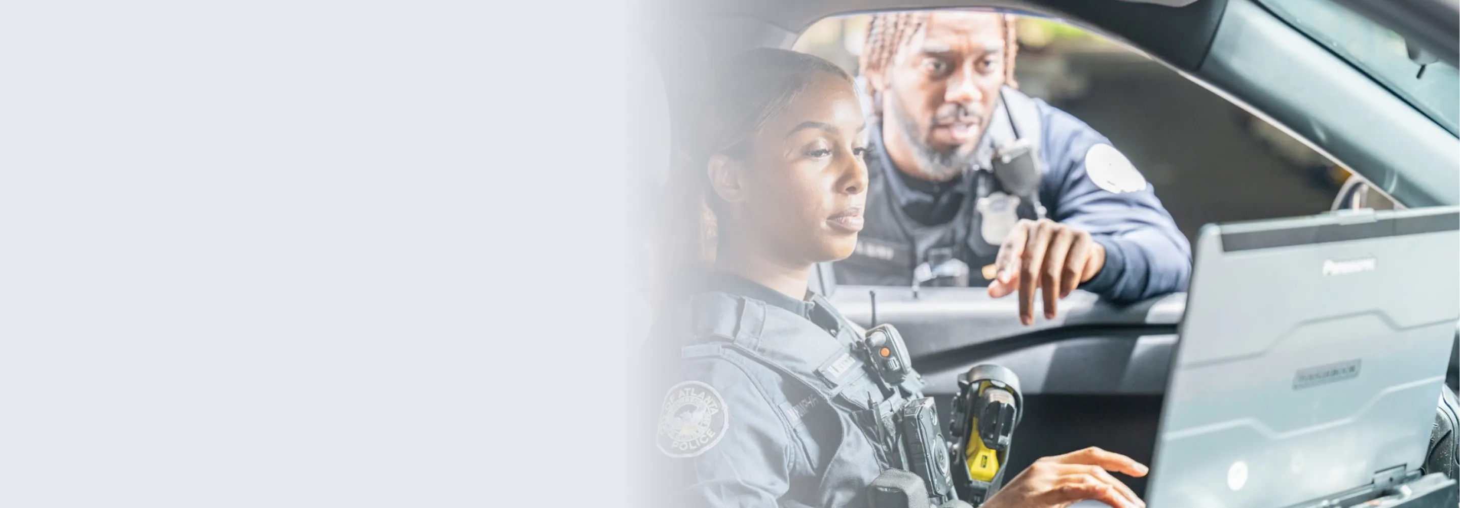 Two police officers in uniform looking at a laptop inside a patrol car, with one officer seated and the other leaning in to point at the screen.