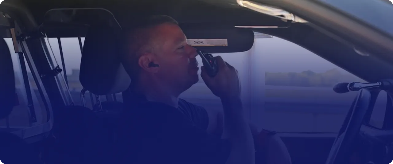 Man sitting in the driver's seat of a vehicle speaking into a handheld radio, wearing an earpiece.