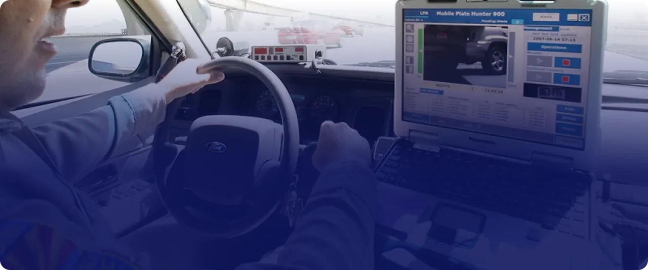 Driver inside a Ford police vehicle using a laptop mounted on the dashboard displaying a license plate recognition software with a vehicle's rear tire and license plate visible on the screen.