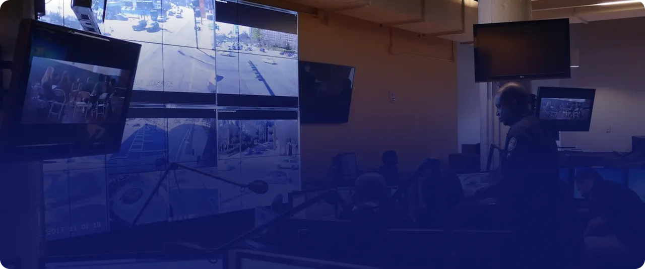 Security control room with multiple large monitors displaying surveillance footage and a person, possibly a security officer, working at a desk nearby.