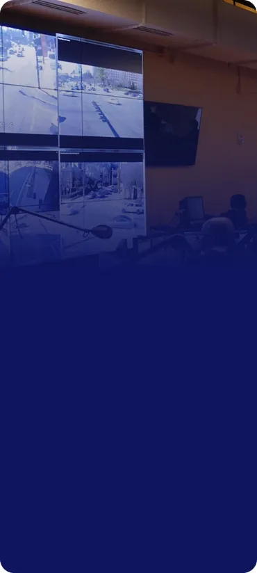 Control room with multiple monitors on the wall displaying live security camera footage of outdoor urban streets and intersections, with people working at desks below.