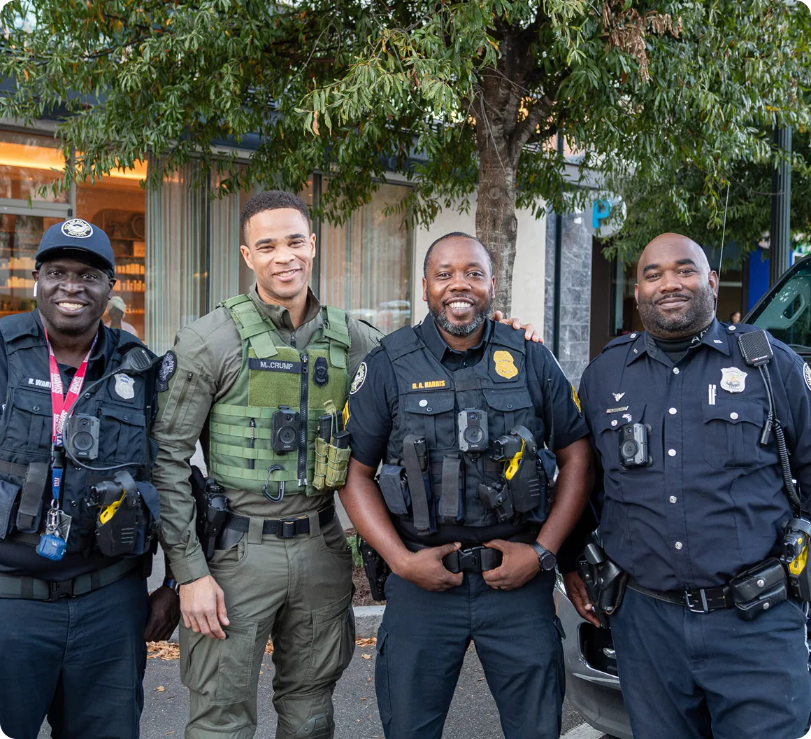 Four smiling police officers standing outside near a tree and a building, wearing uniforms and body cameras, looking at the camera.