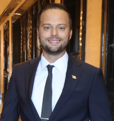 Smiling man wearing a dark suit, white shirt, and black tie with an American flag pin on his lapel, standing indoors with warm lighting in the background.