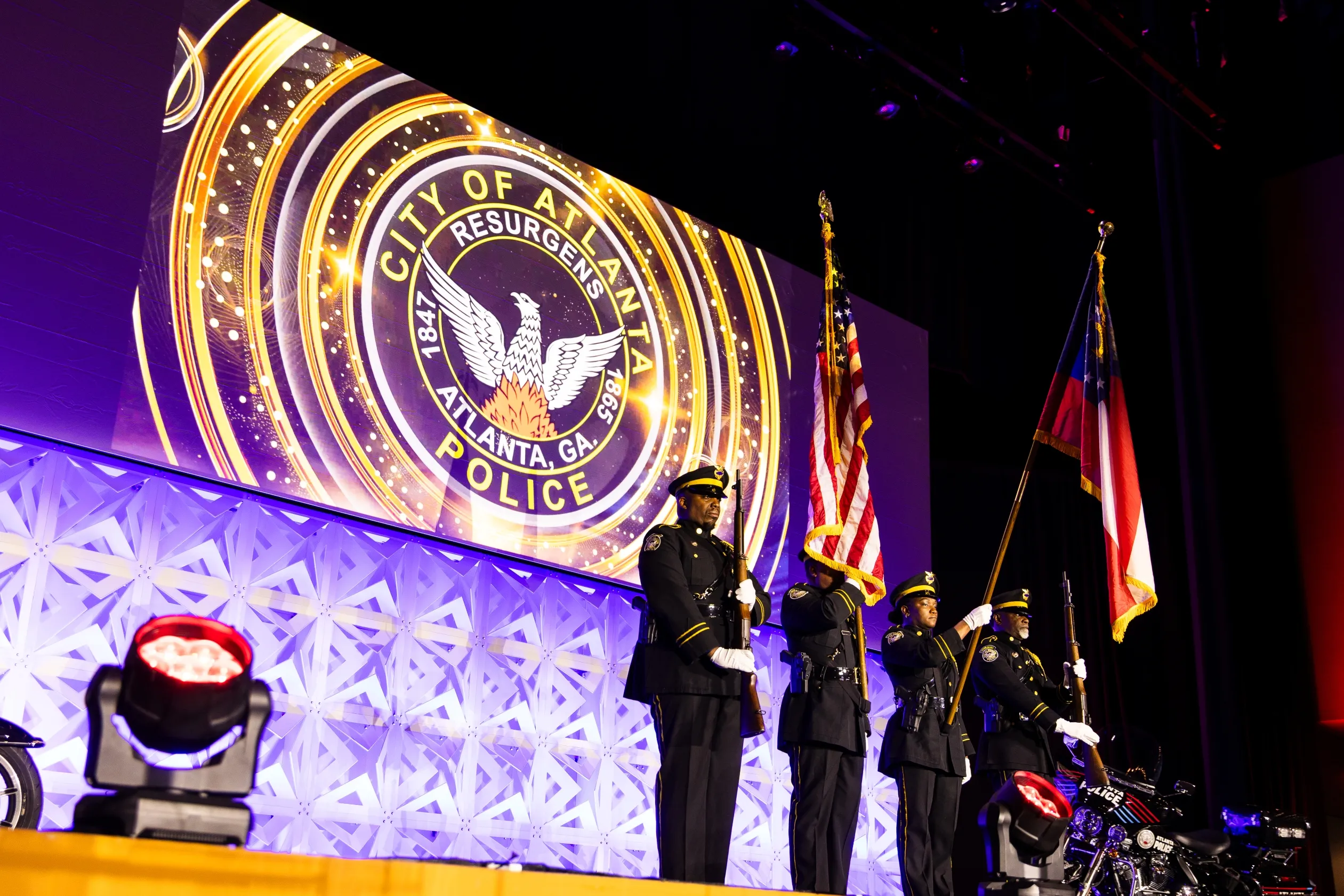 Four uniformed Atlanta police officers standing in formation on stage, holding rifles and flags, with a large illuminated City of Atlanta Police emblem displayed on a screen behind them.