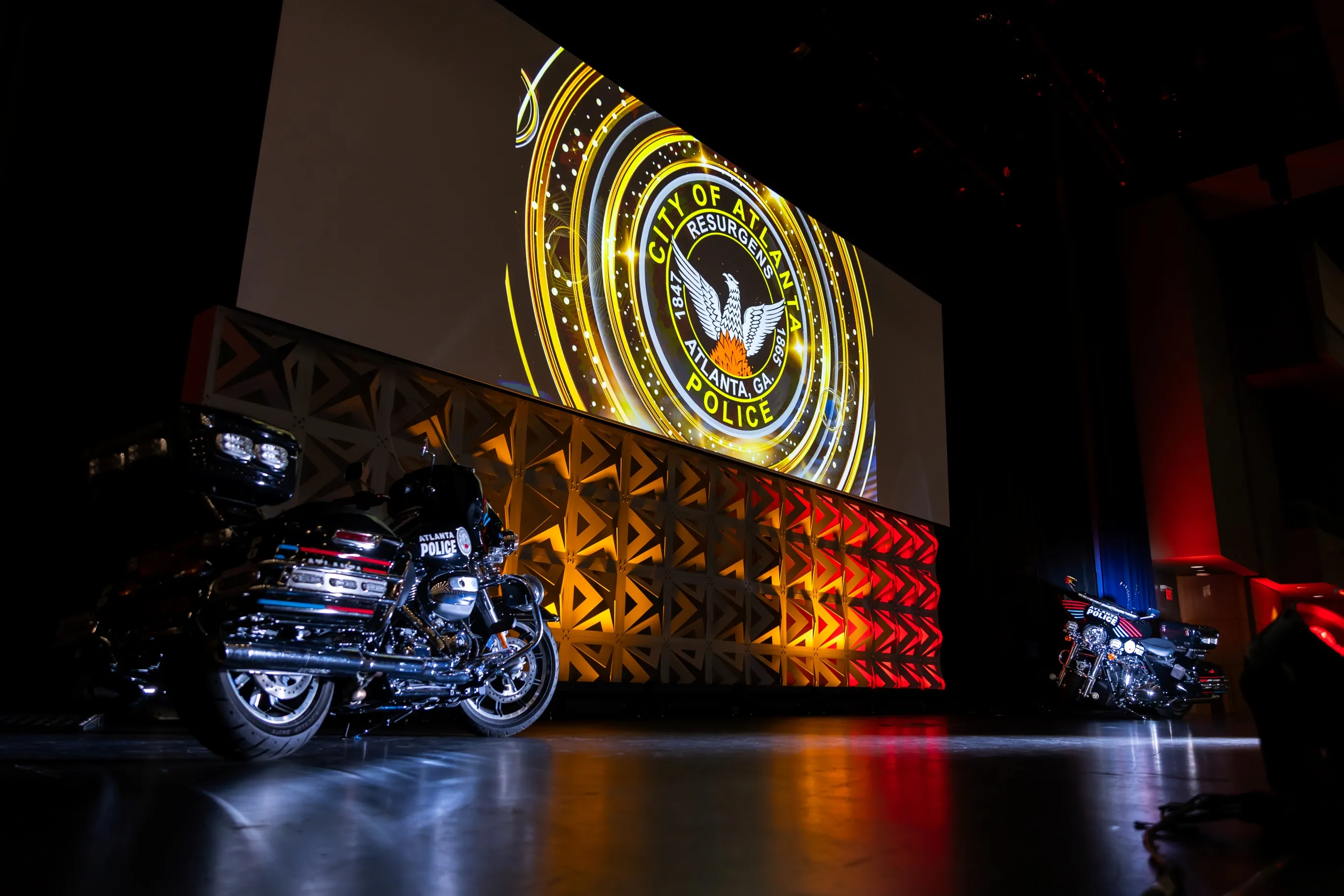 Two black Atlanta Police motorcycles parked on a stage with a large illuminated emblem of the City of Atlanta Police displayed on a screen above a geometric patterned wall lit in yellow and red.