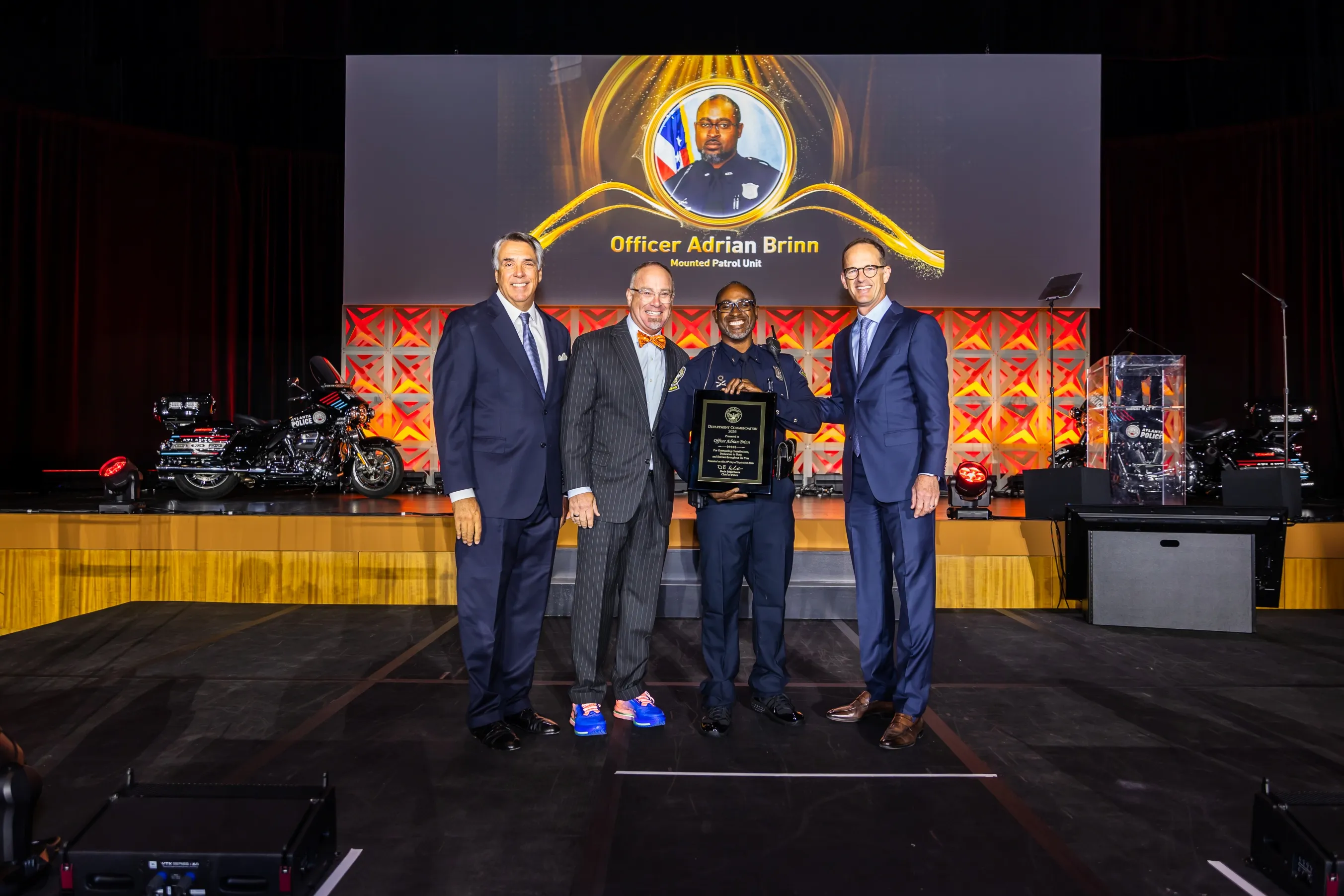 Four men stand on a stage with two police motorcycles in the background; the man second from right, dressed in police uniform, holds a plaque. Behind them is a large screen displaying a photo of Officer Adrian Brinn with his name and Mounted Patrol Unit.