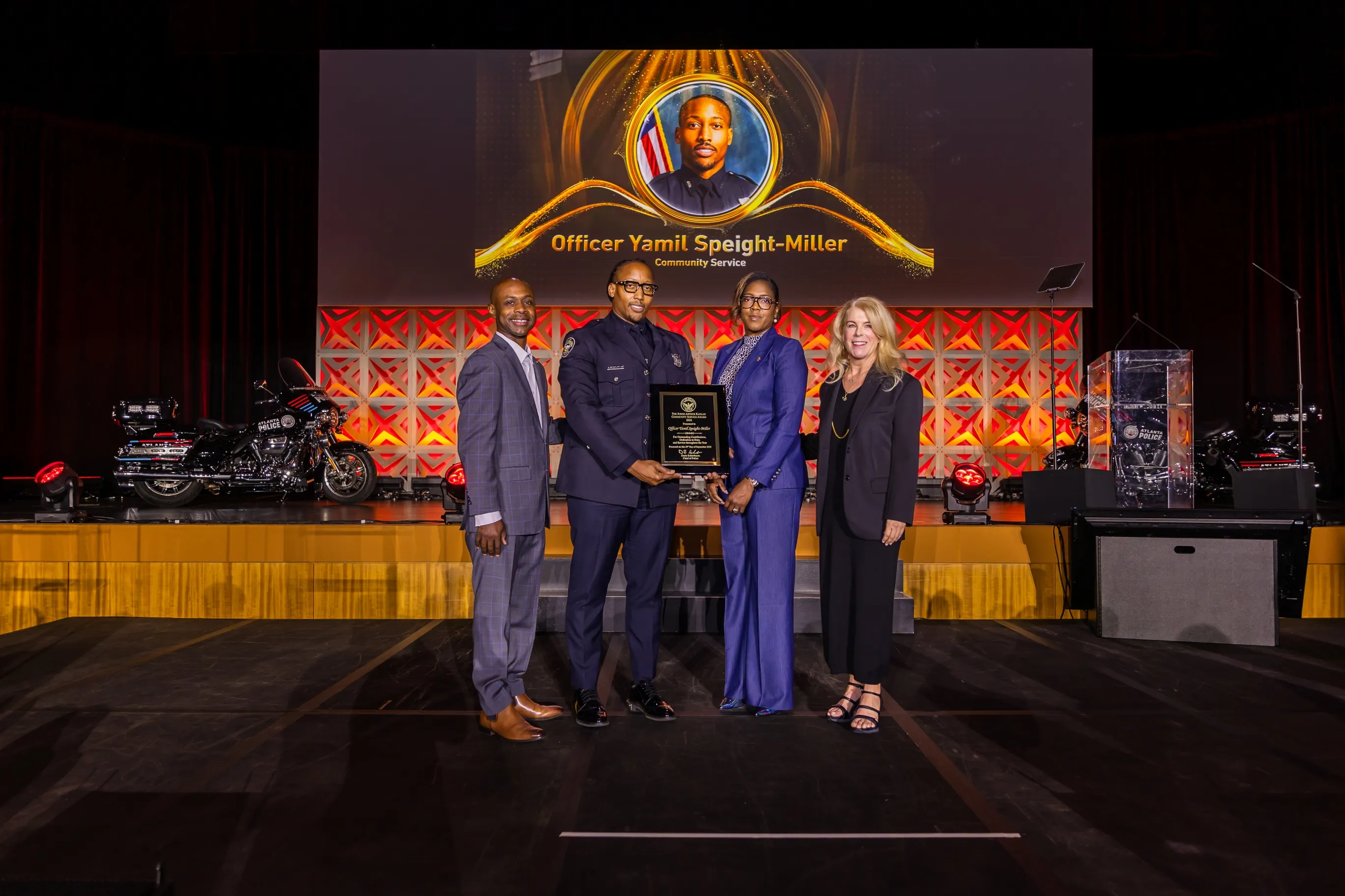 Four people standing on stage in front of a backdrop with a portrait and the name Officer Yamil Speight-Miller, Community Service. One person in a police uniform holds a plaque, flanked by two men and one woman in formal attire, with police motorcycles visible on stage sides.