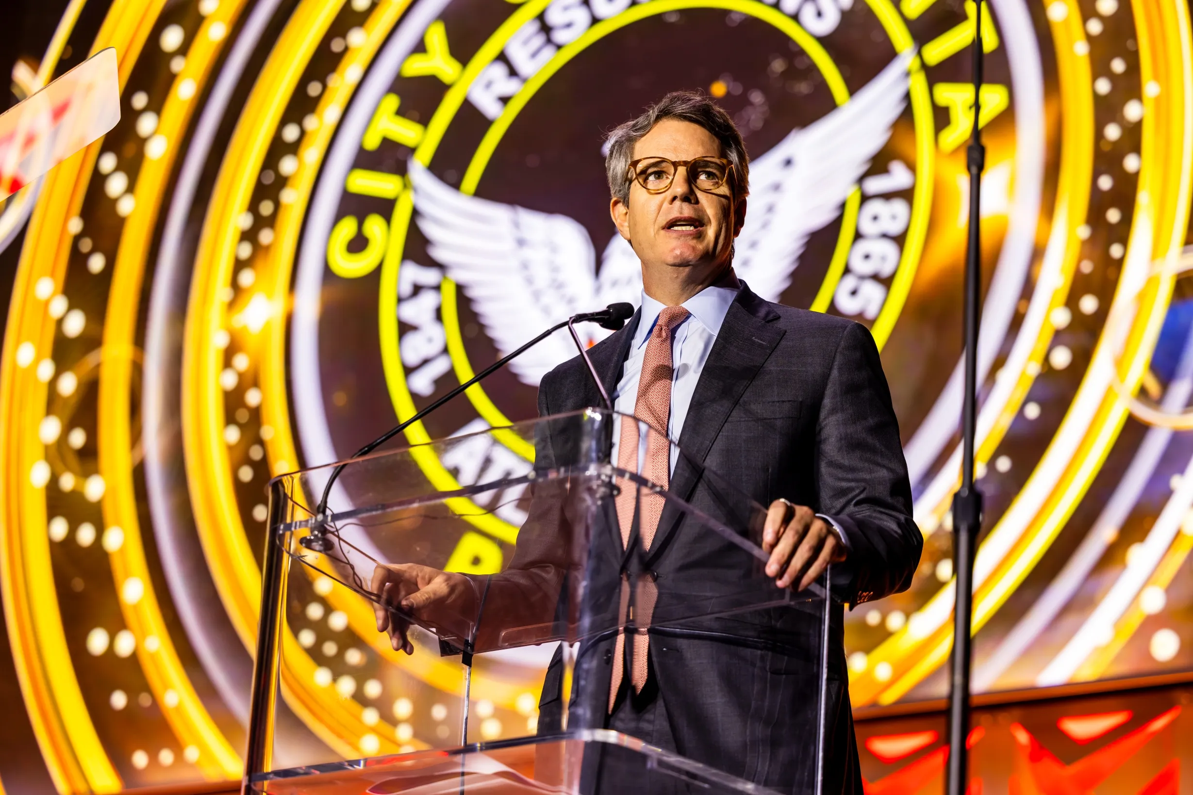 A man in a suit and glasses speaks at a clear podium with a microphone, standing in front of a large, illuminated circular emblem background with text and a white winged symbol.
