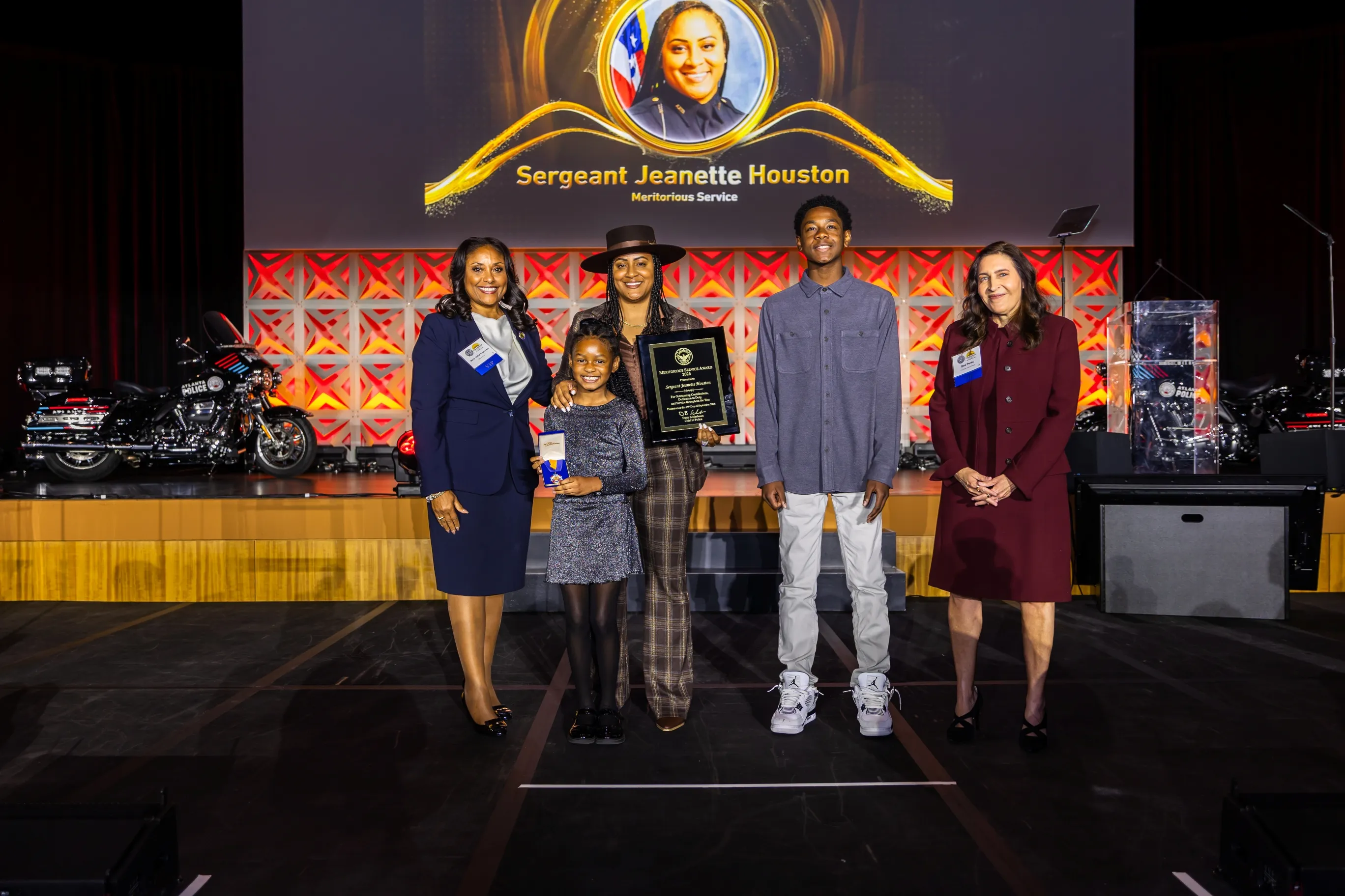 Five people stand on stage in front of a screen showing a portrait and text honoring Sergeant Jeanette Houston for Meritorious Service; one woman in plaid pants holds an award plaque, a girl beside her holds a medal, and two women and a boy stand alongside them with smiles.