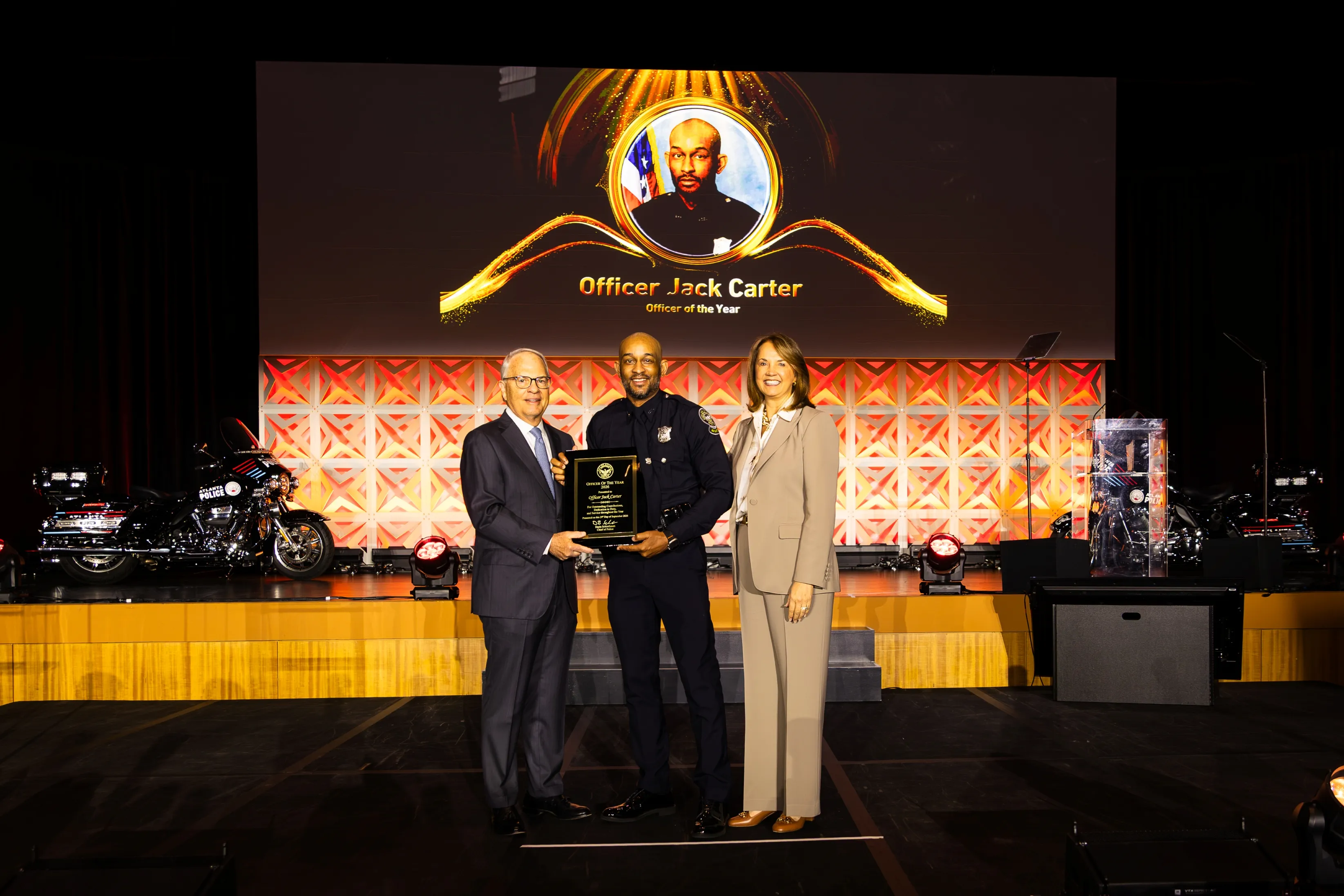 Officer Jack Carter in police uniform holding an award plaque on stage between a man in a suit and a woman in a beige suit, with a backdrop displaying his photo and the title 'Officer Jack Carter Officer of the Year.'
