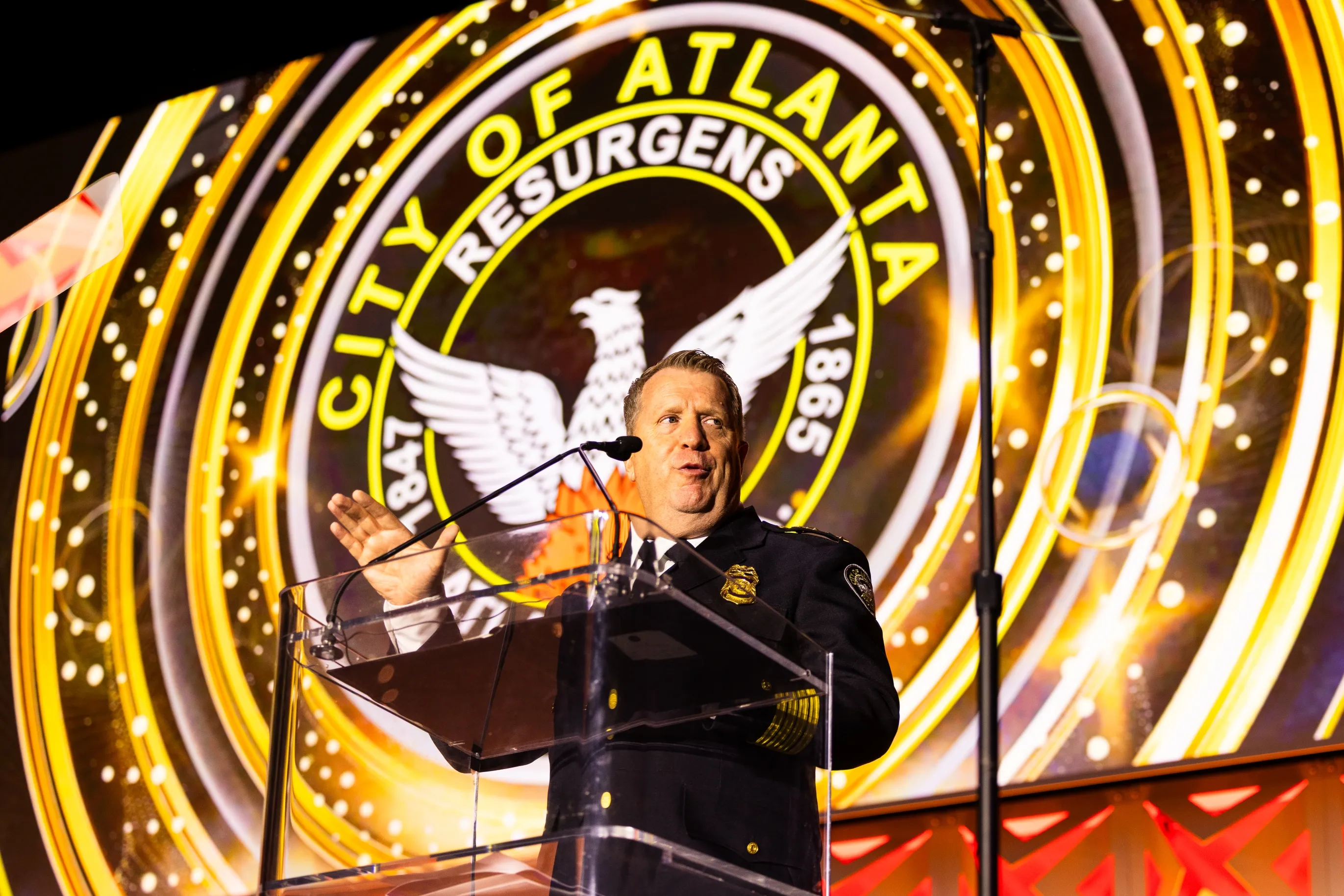 A man in a police uniform speaking at a clear podium in front of a large circular emblem with the text 'City of Atlanta' and an eagle design, illuminated with bright yellow and gold lights.
