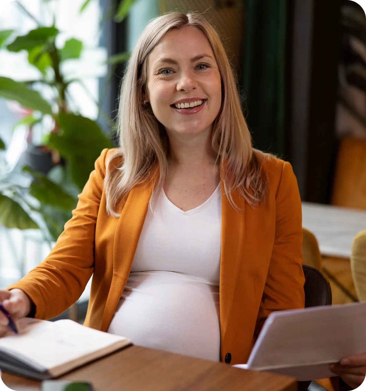 Lächelnde schwangere Frau in einem orangefarbenen Blazer, die an einem Schreibtisch in einem Innenraum Papiere und einen Stift in der Hand hält.