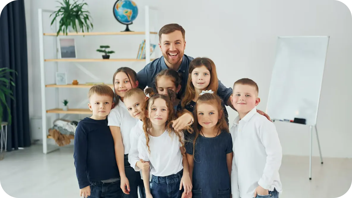 Lächelnder Lehrer, der hinter einer Gruppe von acht Kindern steht, die in einem hellen Klassenzimmer mit einem Globus und einer Weißwandtafel im Hintergrund posieren.