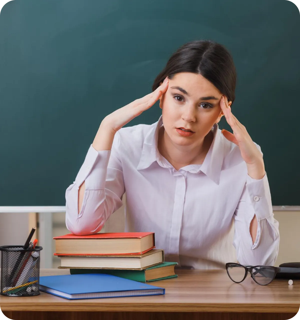 Gestresste Frau sitzt an einem Tisch mit einem Stapel Bücher vor sich, hält ihre Hände an die Schläfen, im Hintergrund eine grüne Tafel.