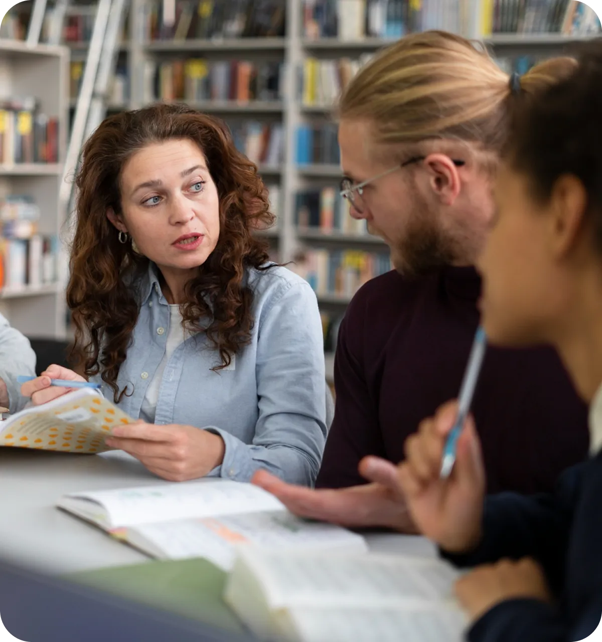 Drei Personen sitzen zusammen an einem Tisch in einer Bibliothek und diskutieren während sie Notizen und Bücher anschauen.