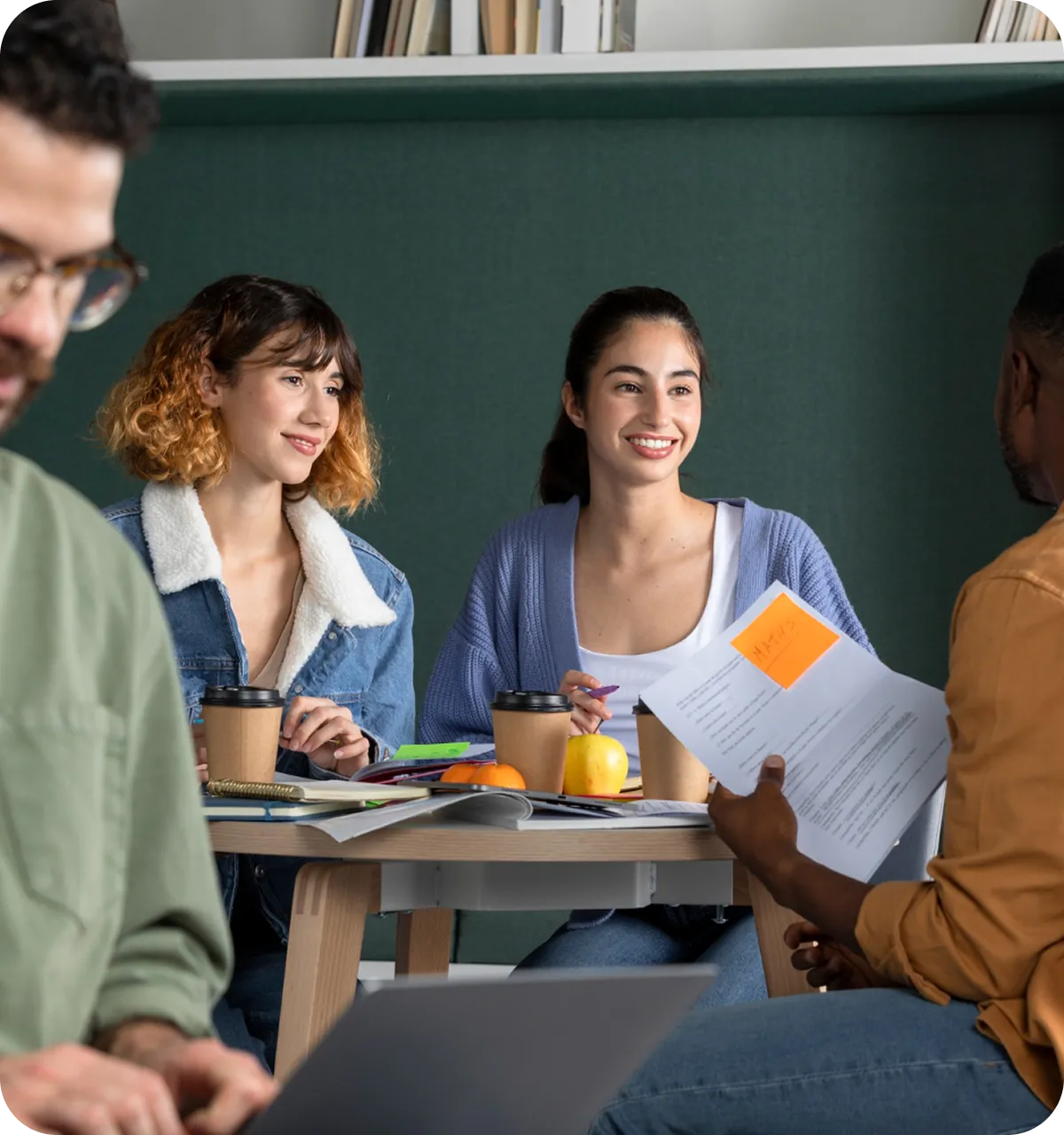 Drei junge Erwachsene sitzen an einem Tisch mit Papieren, Notebook, Kaffee und Obst und unterhalten sich.