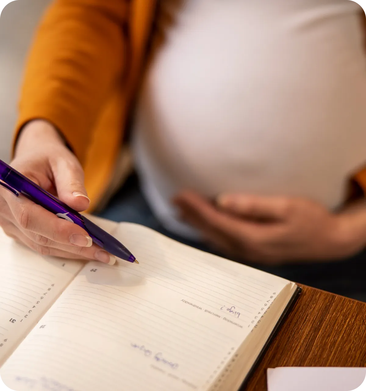 Schwangere Person hält sich den Bauch und hält einen Stift über ein geöffnetes Notizbuch mit Terminen.