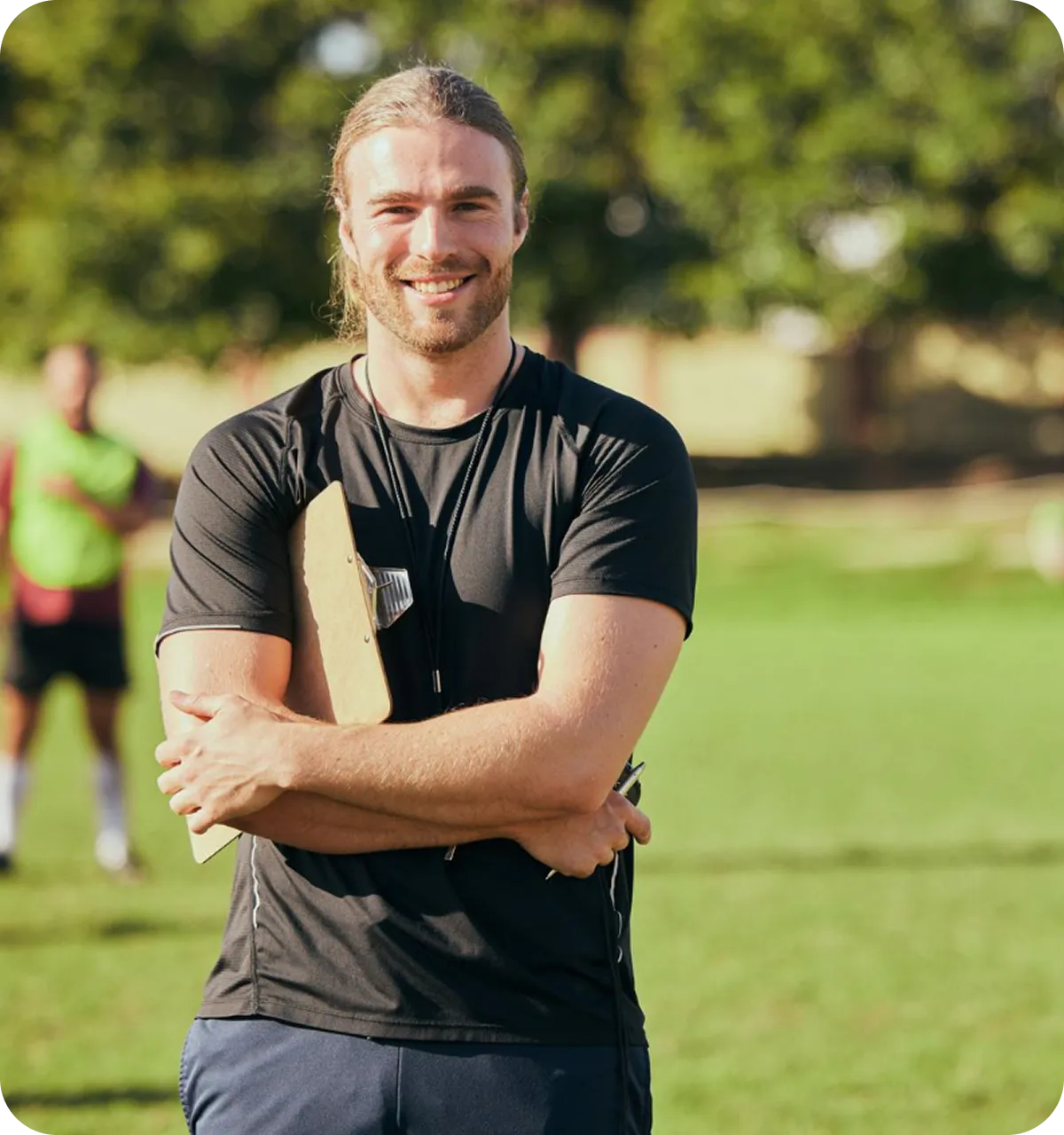 Junger Trainer mit schwarzen Sportkleidung hält ein Klemmbrett und ein Stopper, steht auf einem Sportplatz und lächelt.