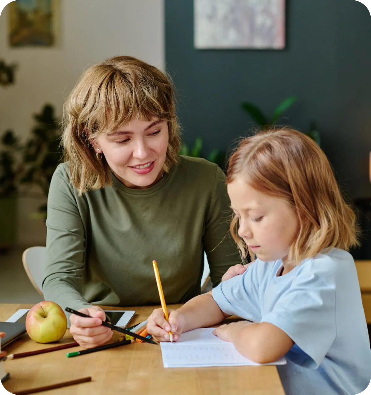 Frau hilft einem Mädchen beim Schreiben in einem Heft an einem Tisch mit Stiften und einem Apfel.
