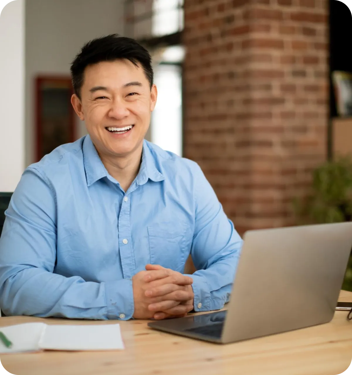 Lächelnder Mann in blauem Hemd sitzt mit verschränkten Händen vor einem Laptop an einem Holztisch im Büro.