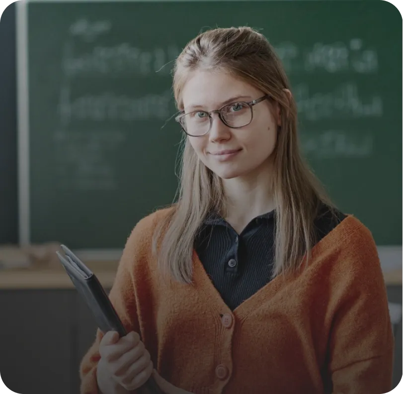 Junge Frau mit Brille und orangem Cardigan hält Unterlagen vor einer Tafel in einem Klassenzimmer.