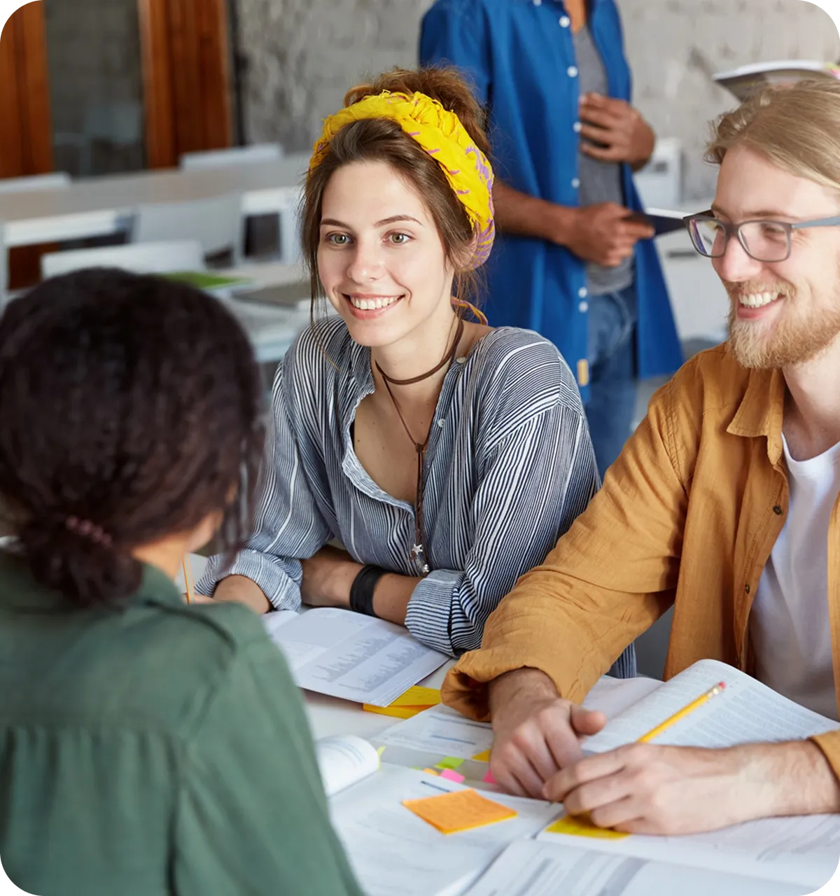 Drei Studierende unterhalten sich fröhlich bei einer Gruppenarbeit mit Büchern und Notizen auf dem Tisch.