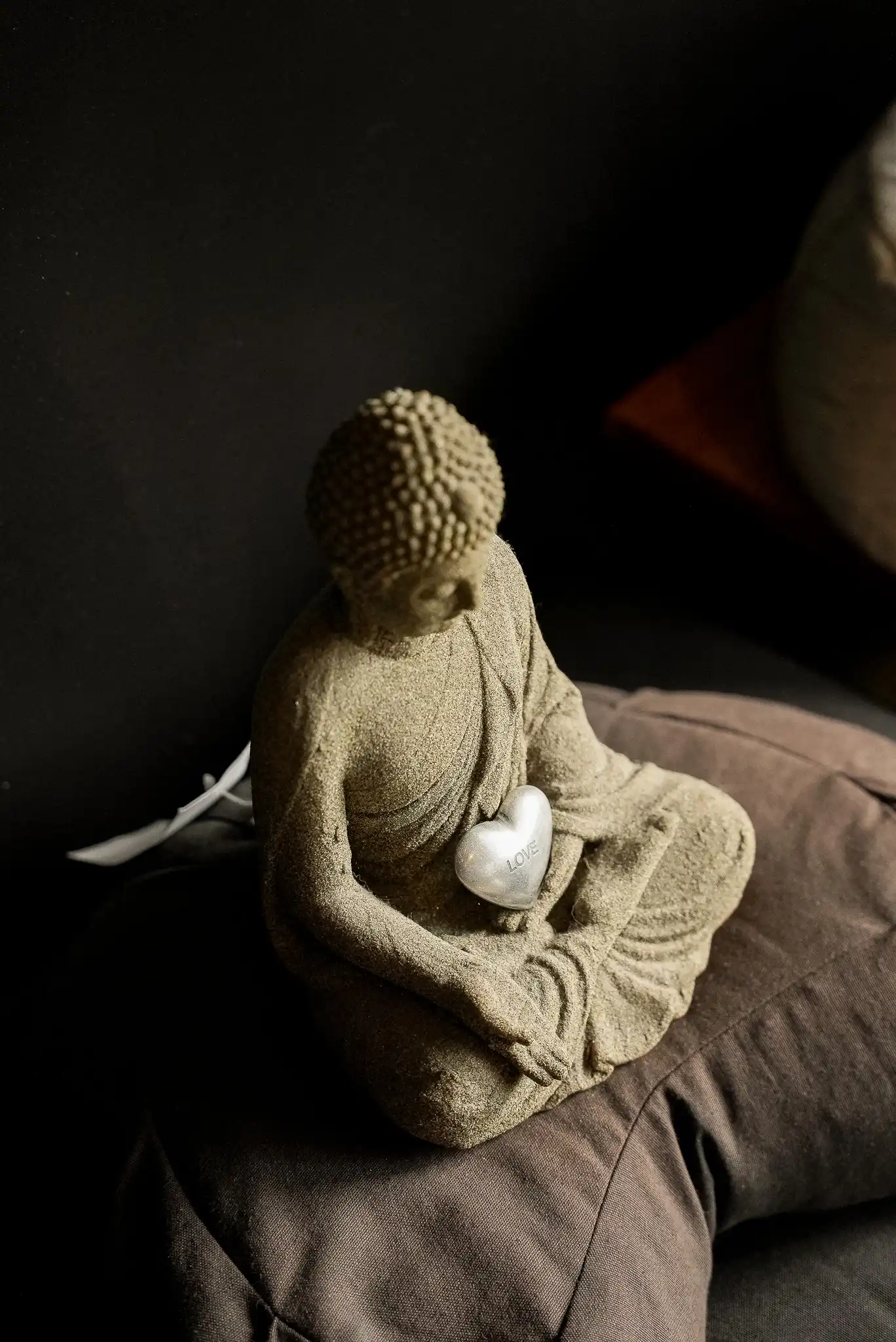 Statuette de Bouddha en pierre tenant un cœur, posée sur des coussins de méditation.