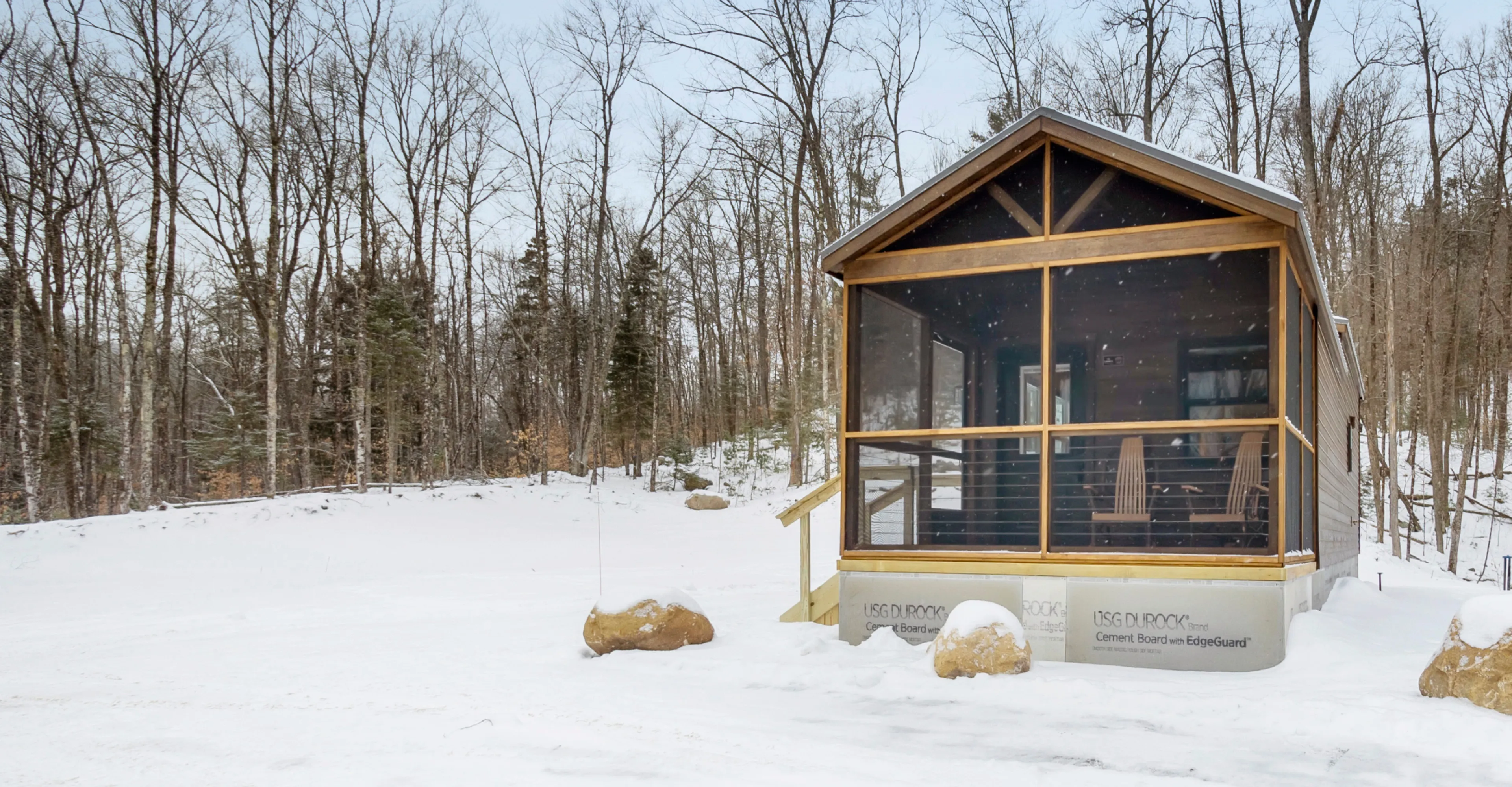 Small wooden cabin with screened porch surrounded by snow and leafless trees in winter.
