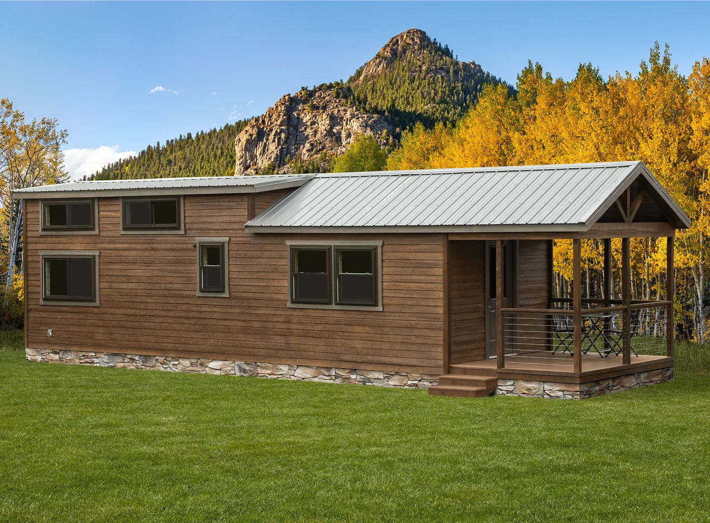 Wooden aspen model cabin with metal roof and front porch set on green grass with autumn trees and rocky mountain in the background.