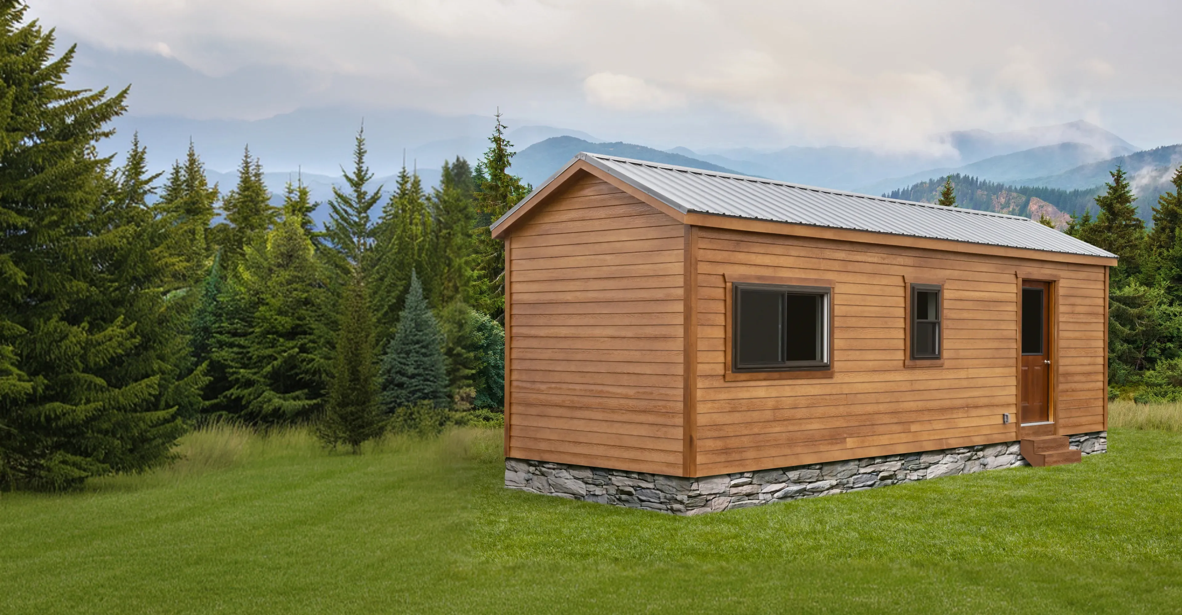 Small wooden cabin with stone foundation on green grass, surrounded by pine trees and misty mountains in the background.