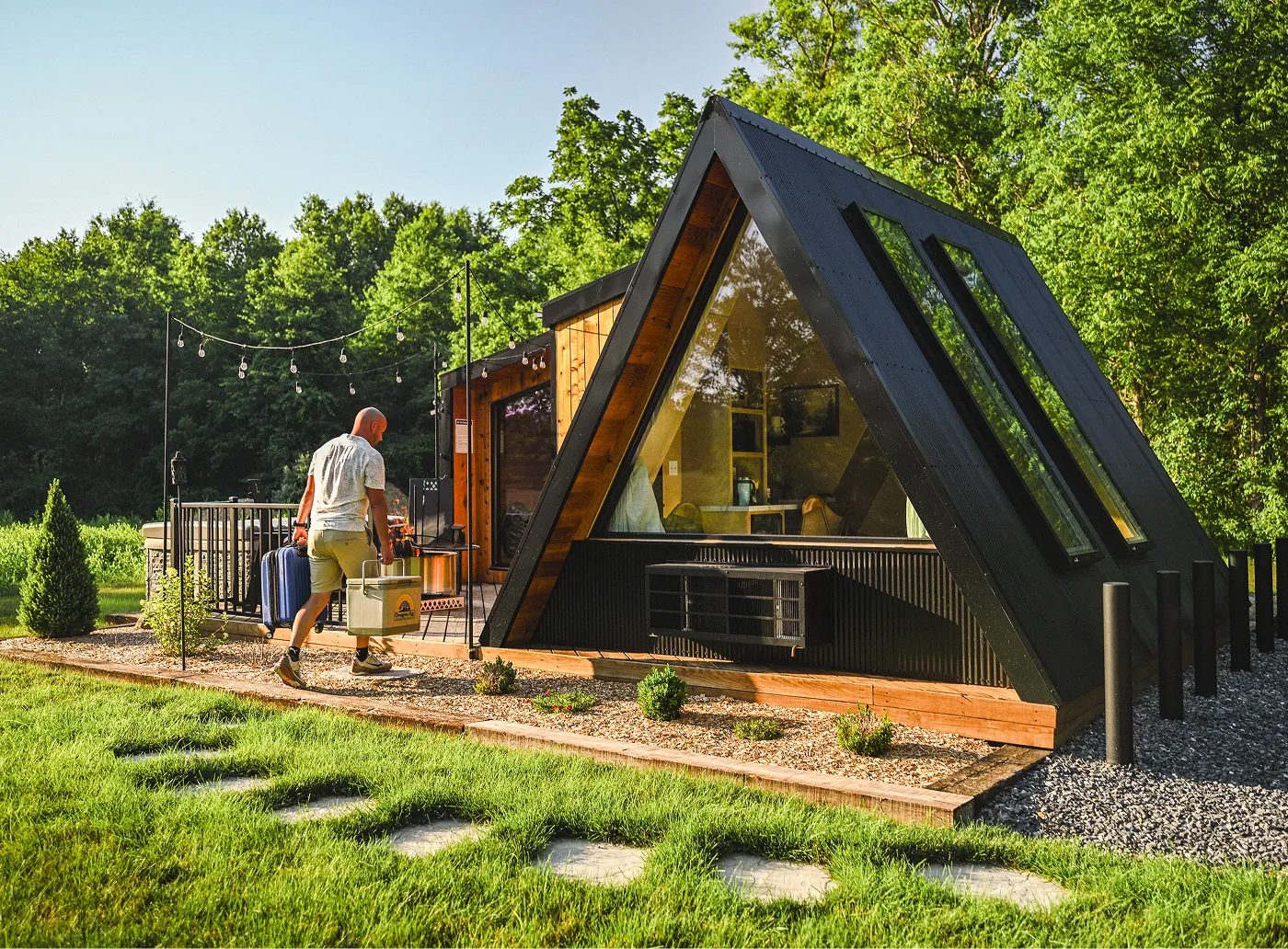 Modern triangular cabin with large windows surrounded by green trees and outdoor string lights.