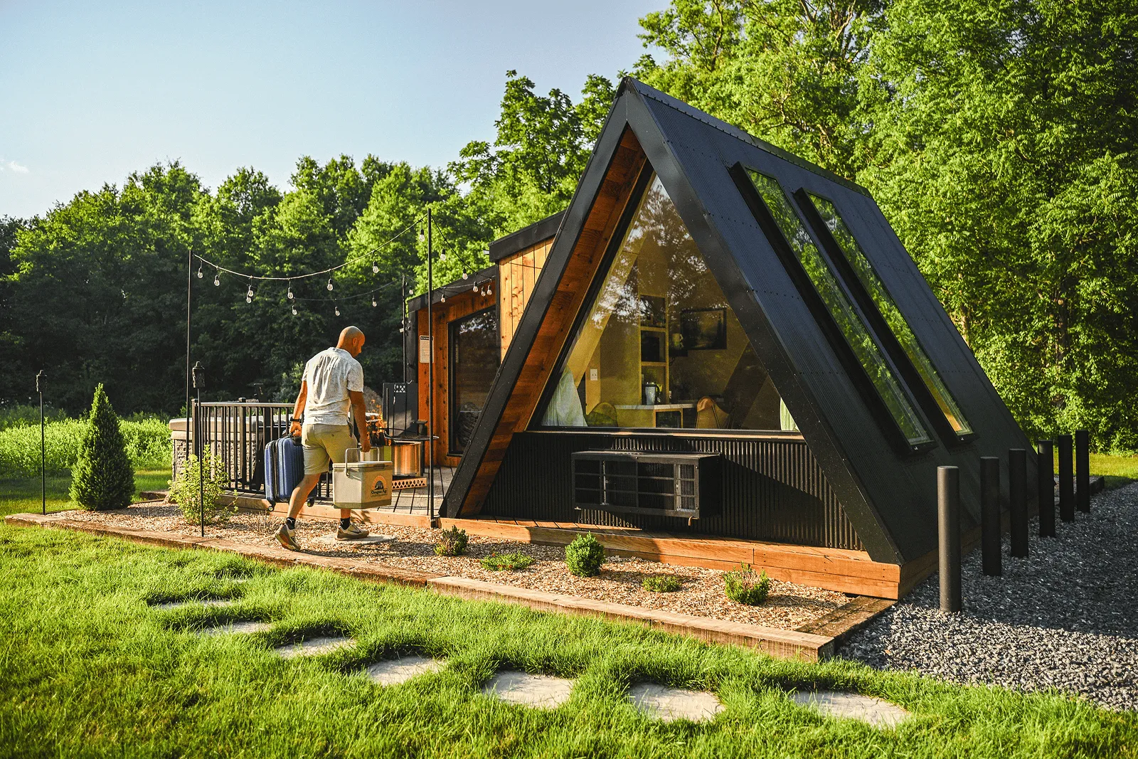 Wooden outdoor deck with two black chairs around a metal fire pit, string lights overhead, and a modern wooden cabin surrounded by greenery.