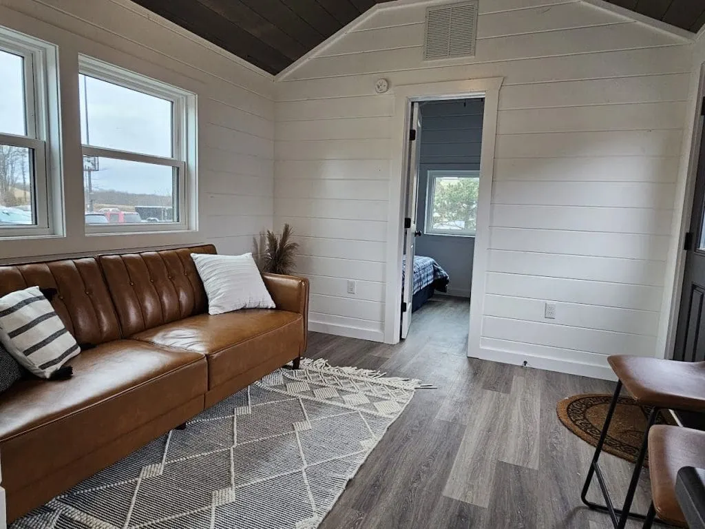 Modern cabin living room with a brown leather couch, patterned rug, two windows, and a view into a bedroom with a bed under a window.